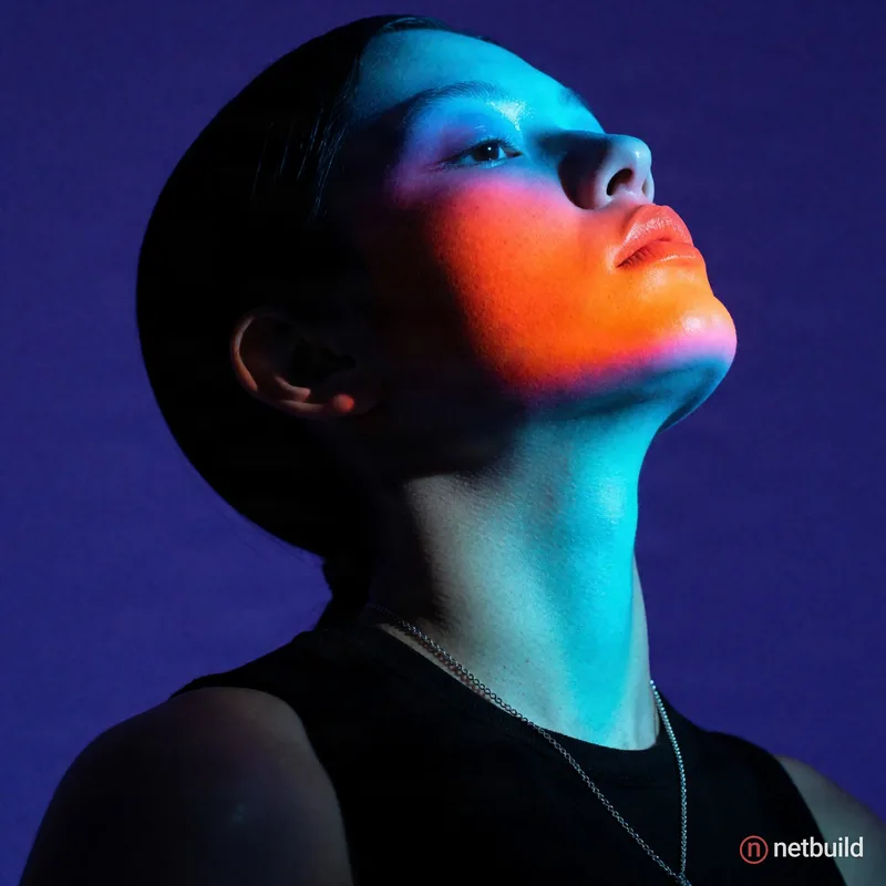 Dramatic low-angle portrait of a figure with head tilted back against a solid indigo background. Deep shadows cloak slicked-back dark hair and a black...